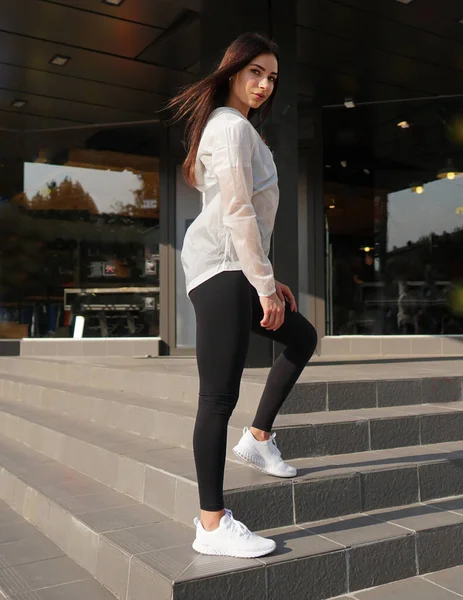 Mağazanın önünde yarım yamalak poz veren çekici genç bir kadın. Beyaz bluz giyen ve antrenman için pantolon giyen bir kız. Modern spor modası kavramı.