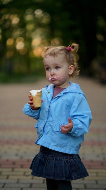 Mavi ceketli, yaz parkında dondurma yiyen küçük bir kız. Çocuk yüzünü dondurmayla boyadı.