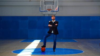 Basketbol sahasında spor ceketli, kot pantolonlu ve ayağının altında basketbol topu olan genç bir adam. Sporcu, kollarını kavuşturmuş, sahanın ortasında duruyor.
