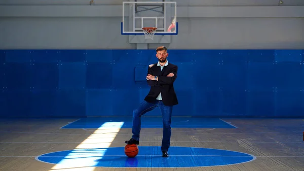 A young man with beard in a blazer and jeans with a basketball under his foot in the game basketball court. Sportsman standing with folded arms in the center of the circle on court