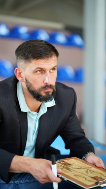 Basketbol sahasında spor ceketli, sakallı genç bir antrenör. Sporcular antrenörlük tabletinde resim çiziyorlar.