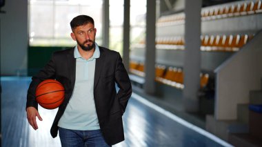 Basketbol sahasında spor ceketli sakallı genç bir adam. Kolunun altında topla duran sporcu