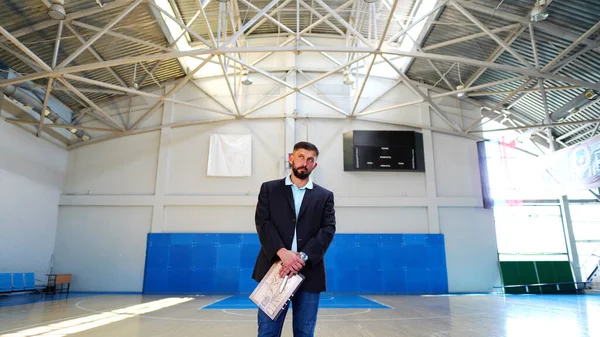 A young man with beard in a blazer in game basketball court. Man standing alone in middle of court with coach tablet