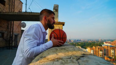 Şafak vakti binanın çatısında basketbol tişörtlü, orta yaşlı sakallı bir adam güneşli bir şehrin arka planında. Koç uyanık bir şehre bakıyor.
