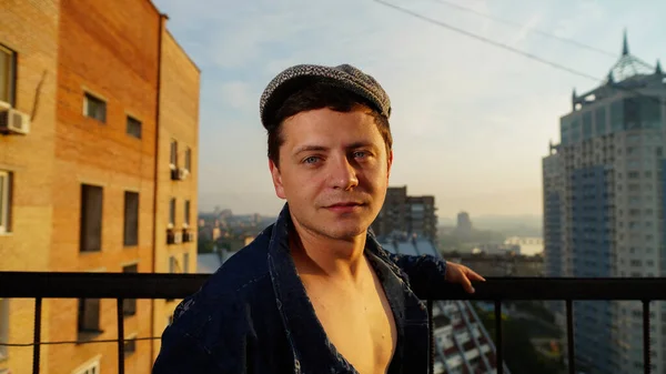 A smart guy in a unbuttoned shirt and flat cap standing on the roof of the building at the dawn against the background of a city rooftops. Man holding railing and looking at the camera