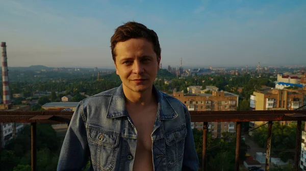 A smart guy in a unbuttoned shirt and flat cap standing on the roof of the building at the dawn against the background of a city rooftops.