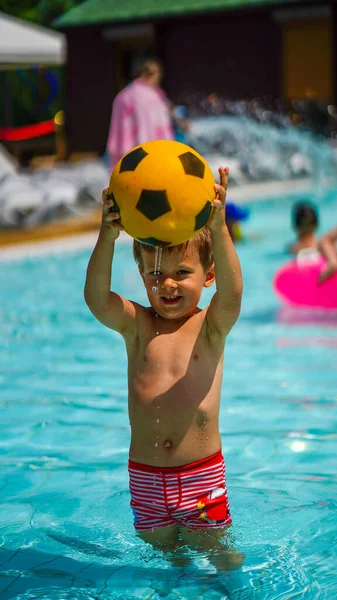 小さなかわいい男の子立って膝深いで澄んだ水保持黄色ボールのためにサッカーのために頭の上にウォーターパーク — ストック写真