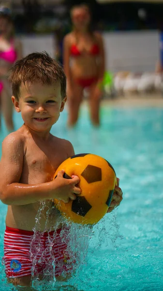 水公園でサッカーのための黄色のボールを保持する澄んだ水の中に膝の深立ってかわいい男の子 — ストック写真