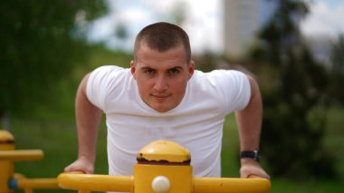 Beyaz tişörtlü güçlü genç adam, dayanıklı eğitim ekipmanlarıyla çalışıyor. Paralel çubuklarda yukarı iten adam.