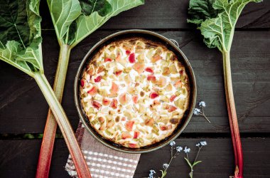 Koyu kahverengi ahşap masa zemininde kaymak ve ekşi kremalı ev yapımı yuvarlak ravent (Rheum hybridum) turtası. Rhubarb sapı yana yatıyor. Mevsimlik bahar tatlısı.