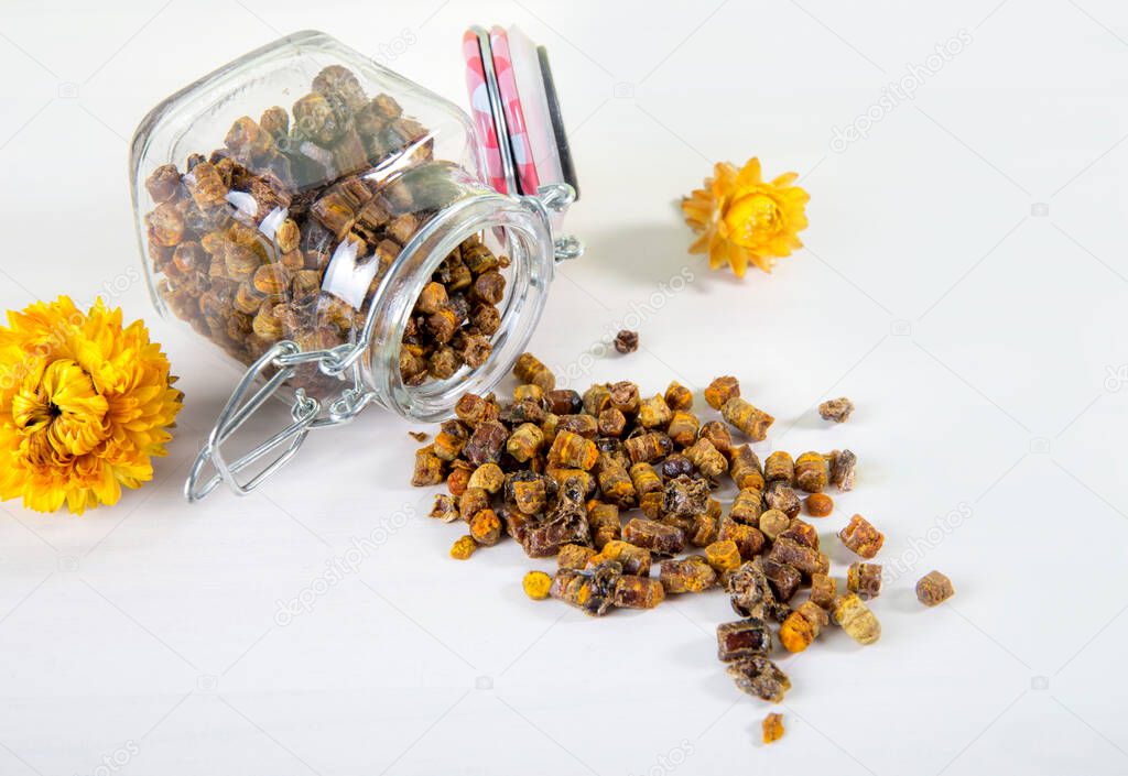  Alternative medicine, domestic honey bee made bee bread ( fermented flower and plant pollen ) inside small glass jar and scattered on white table. Flat lay view.