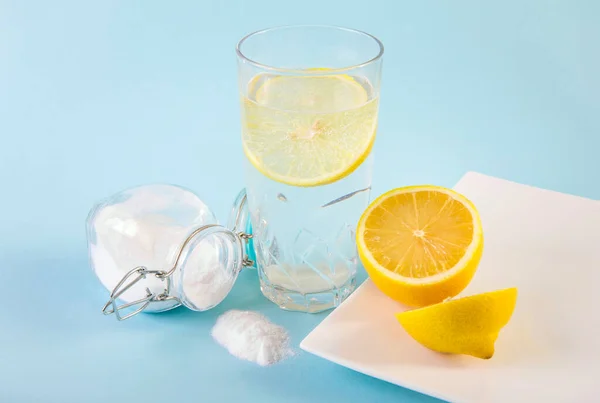 Baking soda in drinking glass with water and lemon slice juice, health benefits for digestive system concept. Light blue background.