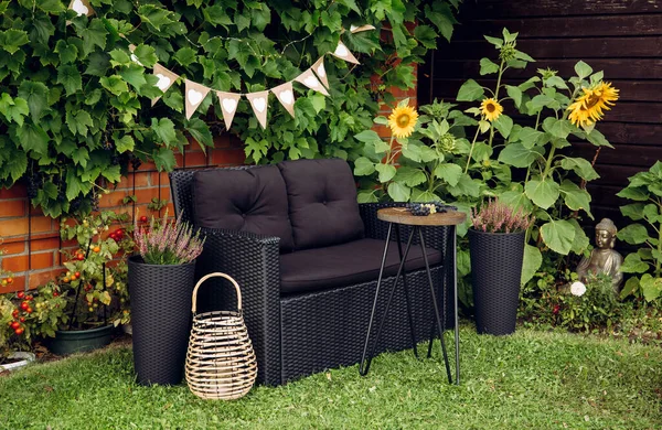 Black modern poly rattan garden furniture chair, metal wire side table. Green grape vines plants growing on red brick wall as decorative element. Wood candle lantern illuminated, pink heather flower.