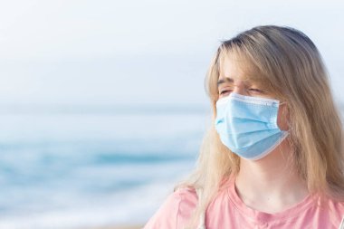 Young woman sad with a blue face mask on the beach in a portrait shot with space for a copyspace wearing a pink t-shirt