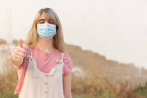 Young Woman Blue Face Mask Background Looking Camera Thumb Wearing — Φωτογραφία Αρχείου
