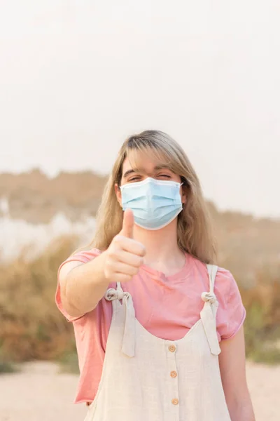 Young Woman Blue Face Mask Background Looking Camera Thumb Wearing — Φωτογραφία Αρχείου