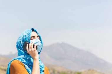Güneşli bir günde cep telefonunda konuşan yüzü maskeli ve tesettürlü genç bir kadının portresi.