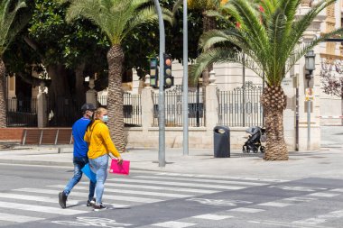 Alicante, İspanya; 30 Haziran 2020; güneşli bir günde, yüzü maskeli bir çift, bir zebra geçidinde karşıdan karşıya geçiyor.