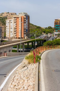 Alicante, İspanya; 8 Temmuz 2020; tramvayın denia caddesinde dolaştığı yeşil yapraklarla dolu köprü, açık gökyüzü olan güneşli bir günde, binalar ve kale ile çevrili.