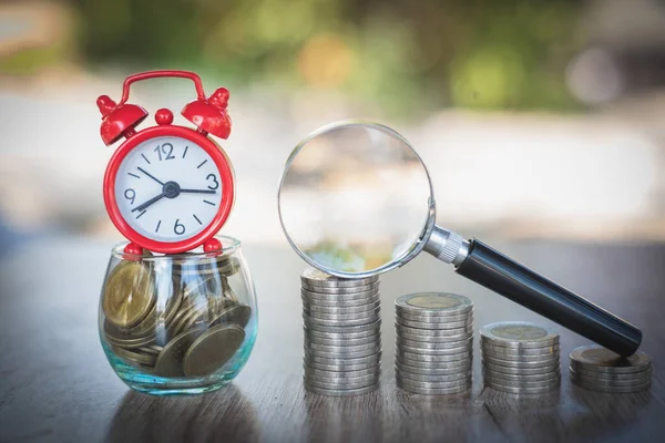 A red alarm clock placed on a glass bottle containing coins. A coin ladder and a magnifying glass.Money saving concept Business growth takes time to invest. Spend time for money Investment risk.