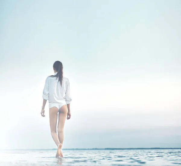 Kaunis seksuaalinen nainen valkoisessa paidassa ja bikineissä kävelemässä rannalla merta ja auringonlaskua vastaan — kuvapankkivalokuva