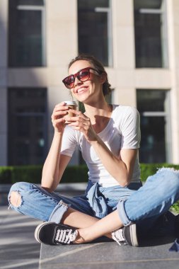 bir genç gülümseyen kadın rahat kıyafetler içinde oturup açık havada kağıt bardak kahve ile onun elinde