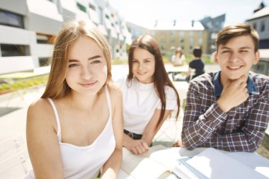 Üç mutlu lise öğrencisinin portresi. Öğrenci arkadaşlar öğrenir, ödevlerini birlikte yapar, zaman geçirir ve eğlenirler. Beyaz gömlekli genç sarışın kız ön planda oturuyor ve kameraya bakıyor.