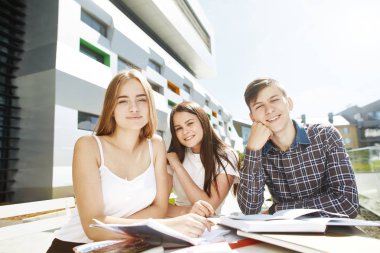 Üç mutlu lise öğrencisinin portresi. Öğrenci arkadaşlar öğrenir, ödevlerini birlikte yapar, zaman geçirir ve eğlenirler. Beyaz gömlekli genç sarışın kız ön planda oturuyor ve kameraya bakıyor.