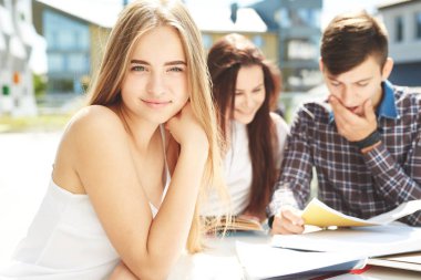 Beyaz tişörtlü genç sarışın kız dışarıda masada arkadaşlarıyla oturuyor ve kameraya bakıyor. Üç mutlu lise öğrencisinin portresi. Öğrenci arkadaşlar öğrenir, ödevlerini birlikte yapar, vakit geçirir ve eğlenirler..