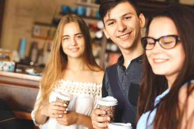 Bir grup arkadaş kafede eğleniyor. Gençler bir kafede buluşuyor. Kâğıt bardaklı genç erkek ve kadınlar kafe tezgahında durup gülümsüyor.