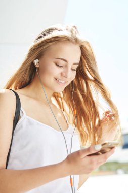 Açık havada kulaklık takan genç sarışın kızın portresi.