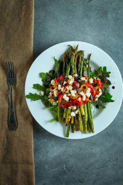 koyu beton arka plan üzerinde beyaz bir tabak üzerinde domates, zeytin, peynir ve arugula ile kavrulmuş kuşkonmaz. masa ayarı, yemek pişirme