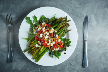 koyu beton arka plan üzerinde beyaz bir tabak üzerinde domates, zeytin, peynir ve arugula ile kavrulmuş kuşkonmaz. masa ayarı, yemek pişirme