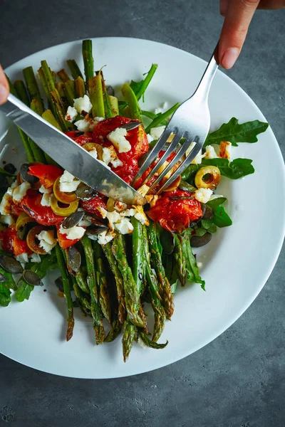 koyu beton arka plan üzerinde beyaz bir tabak üzerinde domates, zeytin, peynir ve arugula ile kavrulmuş kuşkonmaz yeme. kişi çatal ve bıçak ile yemek yiyor. vegan gıda konsepti