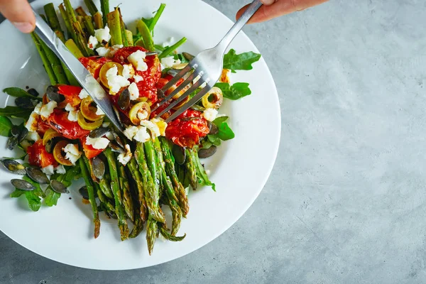 koyu beton arka plan üzerinde beyaz bir tabak üzerinde domates, zeytin, peynir ve arugula ile kavrulmuş kuşkonmaz yeme. kişi çatal ve bıçak ile yemek yiyor. vegan gıda konsepti
