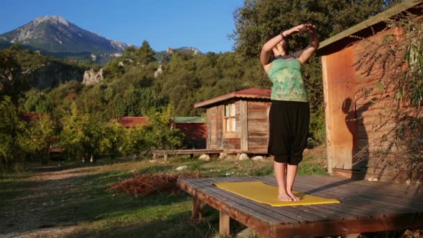 En ung kvinna möter dawn och gör Yoga på verandan på en trä bungalow — Stockvideo