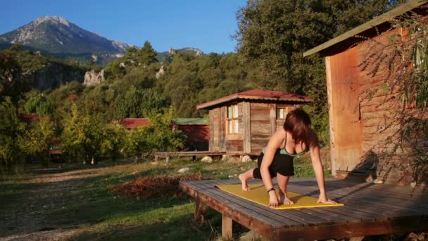 一个年轻的女人遇到黎明, 并在一个木制平房的阳台上做瑜伽 — 图库视频影像