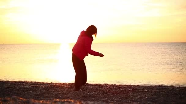 Jovem mulher está encontrando nascer do sol na praia do mar e joga seixos no mar — Vídeo de Stock