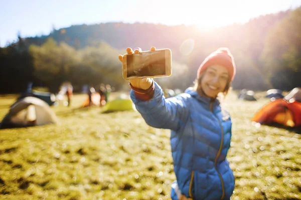 Happy smiling woman wearing in a blue jacket and red cap is standing in campsite in the autumn forest and making selfie on phone camera — Stock Photo, Image