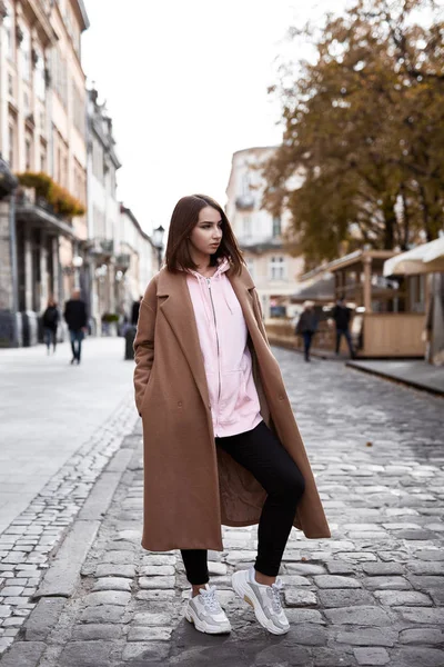 Молодая женщина в коричневом пальто и розовой толстовке позирует на улице — стоковое фото