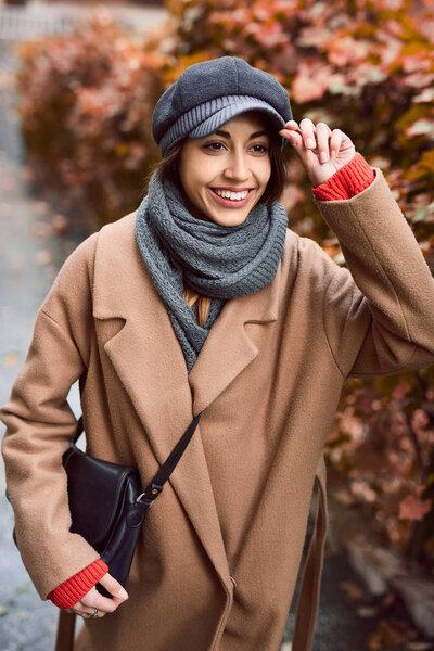 young woman in brown coat and gray cap posing on the street
