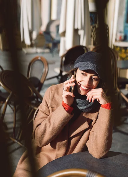 Молодая женщина в коричневом пальто и серой кепке сидит за столом в уличном кафе — стоковое фото