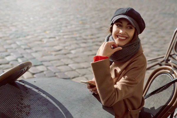 Mujer joven con abrigo marrón y gorra gris está sentado a la mesa en la cafetería de la calle — Foto de Stock
