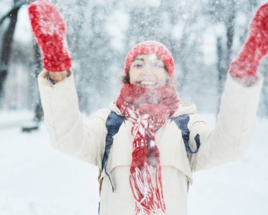 Beyaz aşağı ceket ve kırmızı şapkalı neşeli gülümseyen kadın, atkı ve mittens yürüyüş ve şehirde kar fırtınası sonra karlı sokakta kar ve kartopu plaing. kar fırtınası ve kar yağışı sonrası kış kenti.