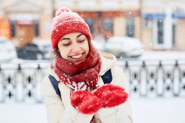 Beyaz aşağı ceket ve kırmızı şapkalı neşeli gülümseyen kadın, atkı ve eldiven karlı sokakta yürüyüş ve şehirde kar fırtınası sonra kar taneleri yakalamak. eldivenlere odaklanmak