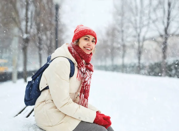 Cheerful smiling woman in white down jacket and red cap, scarf and mittens sitting on a bench in the alley on the snowy street after blizzard in city. winter city after blizzards and snowfall.