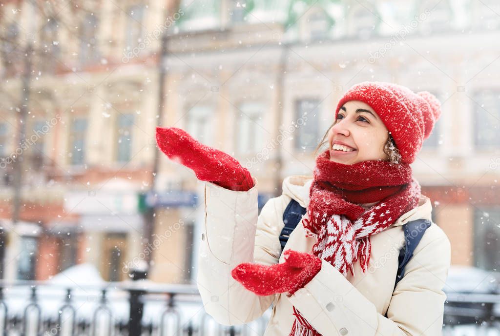 Cheerful smiling woman in white down jacket and red cap, scarf and mittens walking on the snowy street and catching snowflakes after blizzard in city. focus on the mittens