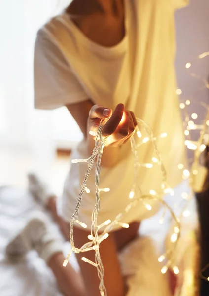 Jente i hvit T-skjorte og shorts, sittende på sengen med hvitt laken og henge et glødende julelys på en bokhylle – stockfoto