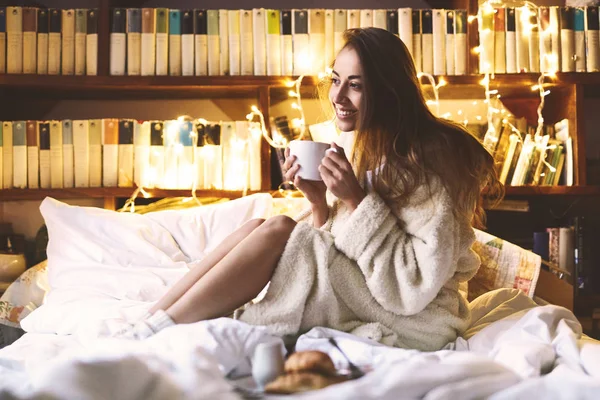 Girl in white warm bathrobe sitting on the bed with glowing christmas garland lights on background and having breakfast — Stock Photo, Image