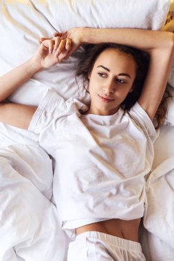 Beyaz bedsheets ile yatakta yatıyordu bir parlak cinsel gülümseyen kız beyaz t-shirt ve şort, üstten görünüm